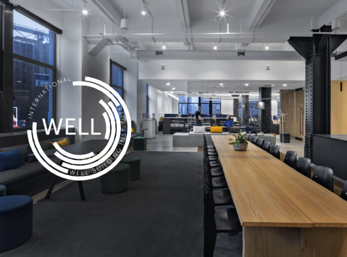 A modern office interior with industrial design elements, a large wooden table, black chairs, and a seating area with a logo of the International WELL Building Institute overlaid on the image.