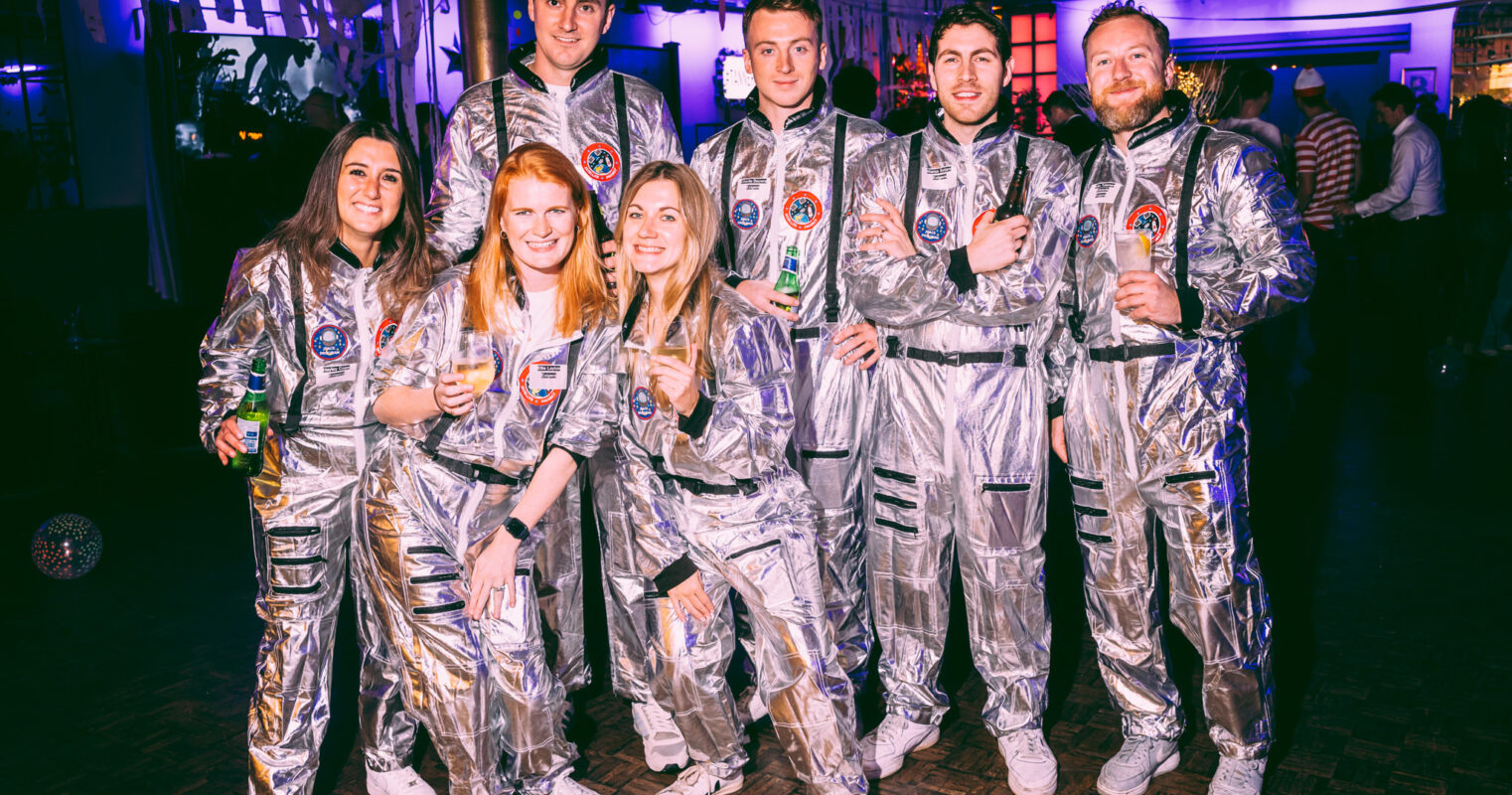 A cheerful group in silver astronaut costumes stands together, holding drinks and smiling.