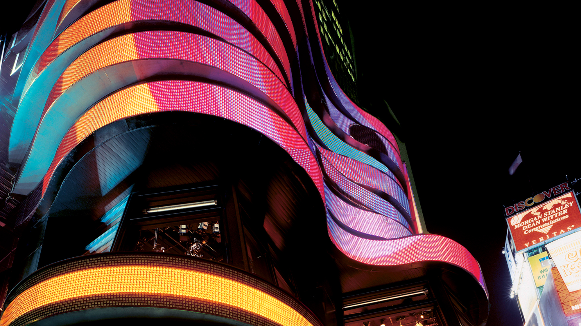 Night view of a dynamic building facade featuring undulating ribbons of LED screens, casting vibrant hues of red, blue, and yellow light. The structure's curves juxtapose against the surrounding urban signage, creating a modern, lively atmosphere.
