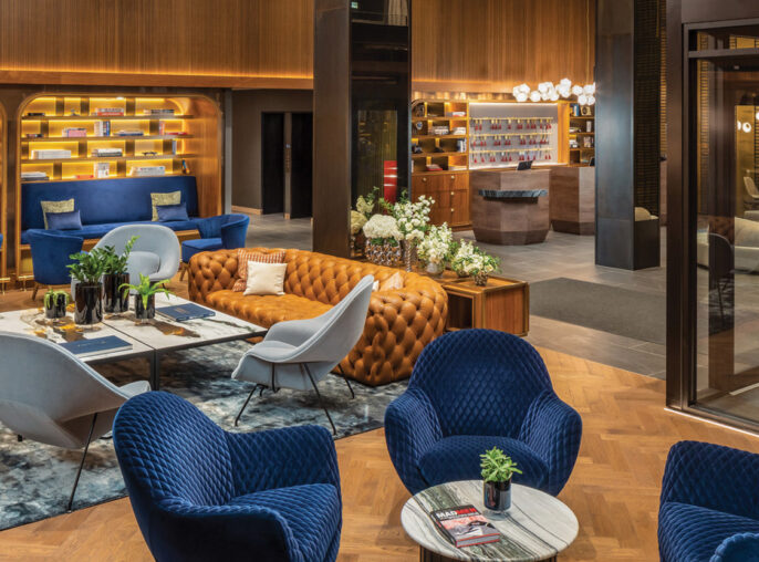 Modern hotel lobby featuring an eclectic mix of furniture styles, with deep blue velvet chairs, caramel button-tufted sofas, and a bold red sculptural seat. A structured, tiered black chandelier hangs overhead, contrasting the warm wooden wall panels and honeycomb-style shelving filled with books.