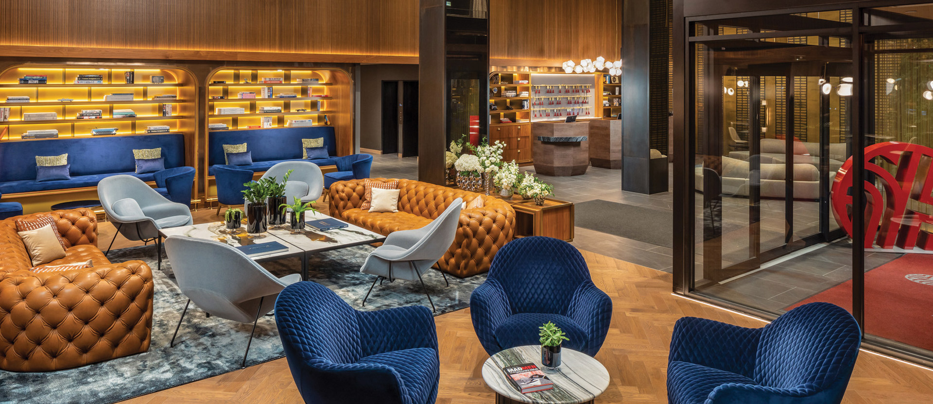 Modern hotel lobby featuring an eclectic mix of furniture styles, with deep blue velvet chairs, caramel button-tufted sofas, and a bold red sculptural seat. A structured, tiered black chandelier hangs overhead, contrasting the warm wooden wall panels and honeycomb-style shelving filled with books.