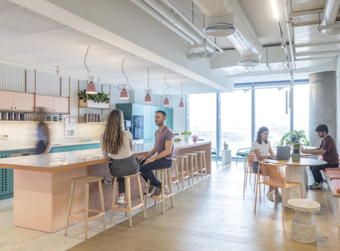 Modern and airy office kitchen and break area with pastel pink and teal cabinetry, terrazzo countertops, and pendant lighting. People engage casually around the bar seating and work at a window-side communal table, highlighting a collaborative atmosphere.