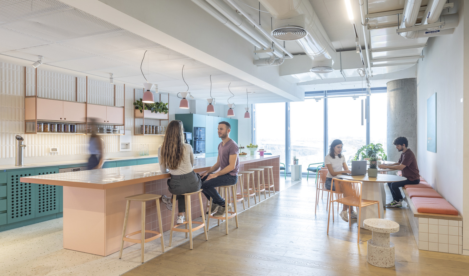 Modern and airy office kitchen and break area with pastel pink and teal cabinetry, terrazzo countertops, and pendant lighting. People engage casually around the bar seating and work at a window-side communal table, highlighting a collaborative atmosphere.