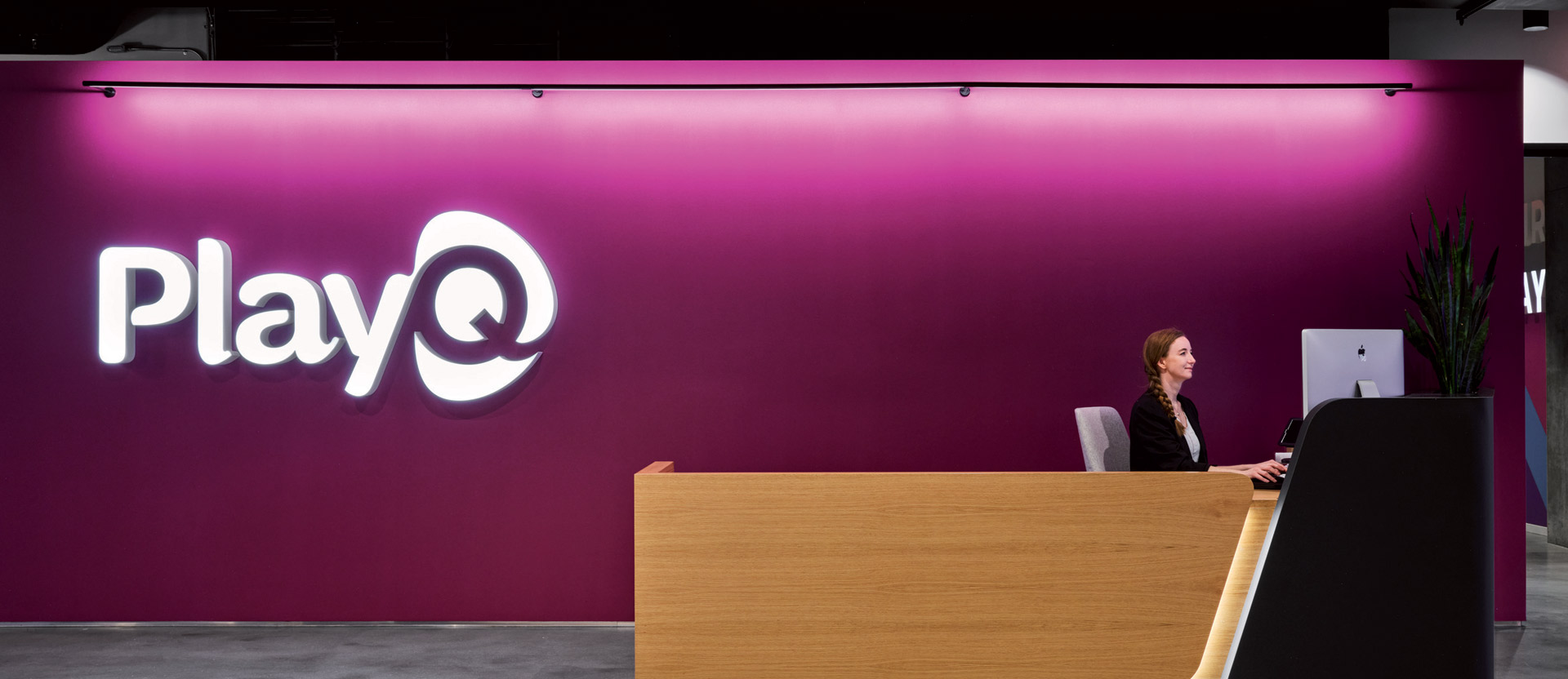 A sleek reception area featuring a wooden desk with a person seated, illuminated by a vibrant pink backlit wall showcasing the 'Play' logo, creating a dynamic corporate ambiance.