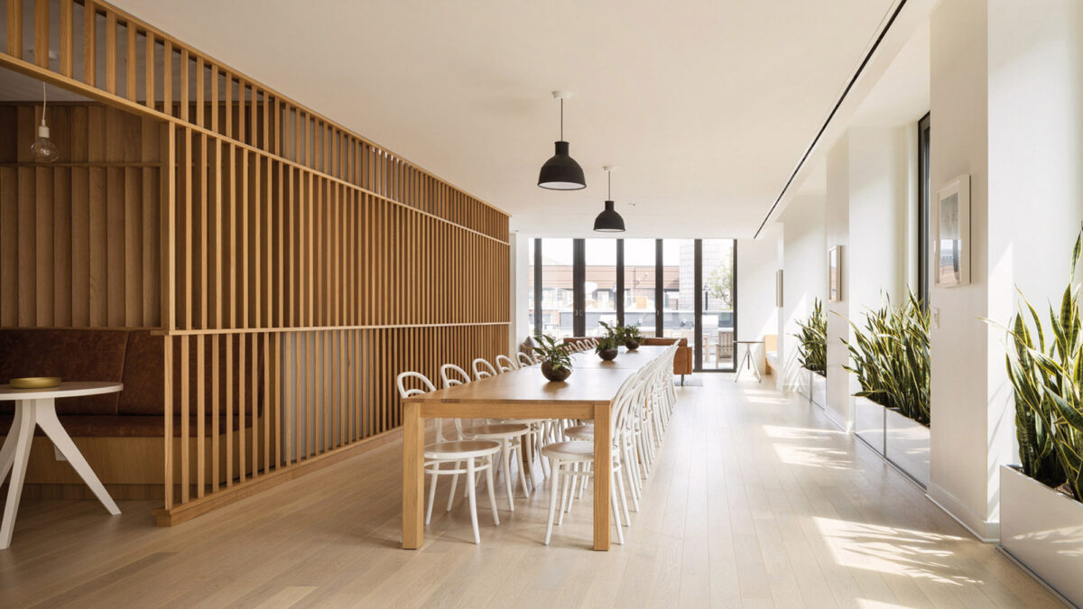 Sun-drenched modern dining space boasting a long wooden table with white chairs, flanked by a sleek wooden slat partition on one side and a gallery-style white wall with framed art on the other. Pendant lights and indoor plants enhance the natural, minimalist aesthetic.