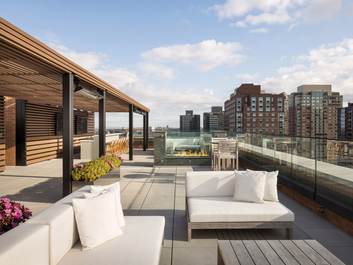 Spacious rooftop terrace with modern outdoor furniture, featuring a white sectional sofa and a sleek fire pit encased in clear glass. The area is framed by wooden pergola structures and glass balustrades, offering a panoramic view of urban skyline and fostering a seamless indoor-outdoor living experience.