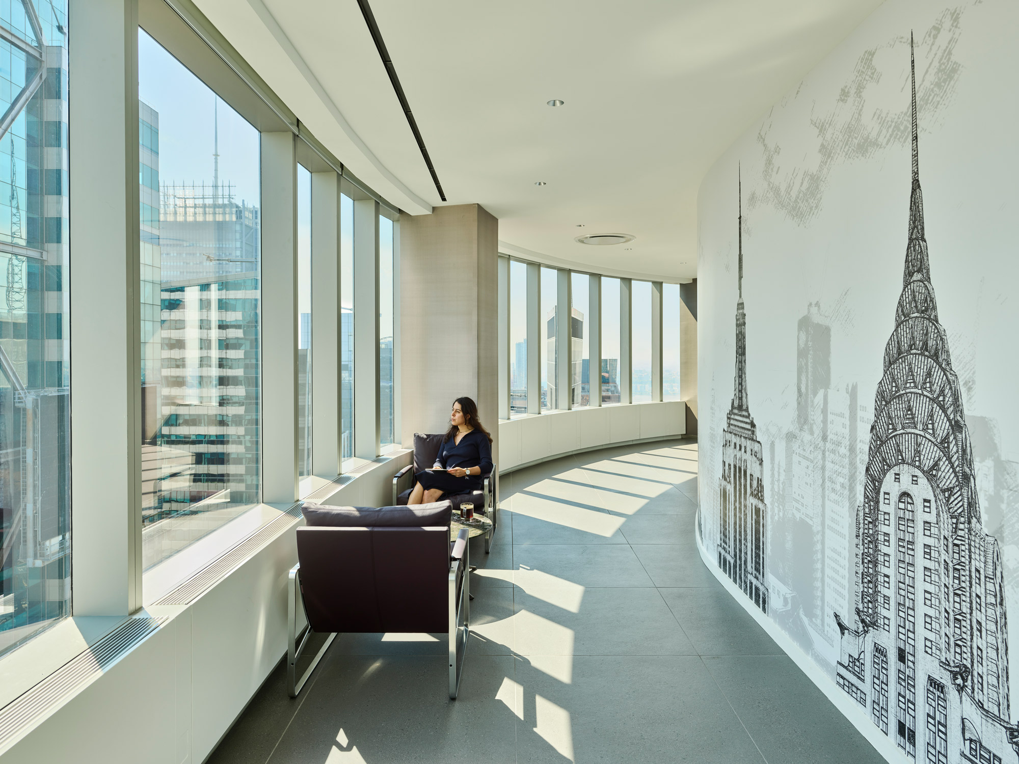 An open office corridor with a seating nook, a large mural of a cityscape, and an employee enjoying the view.