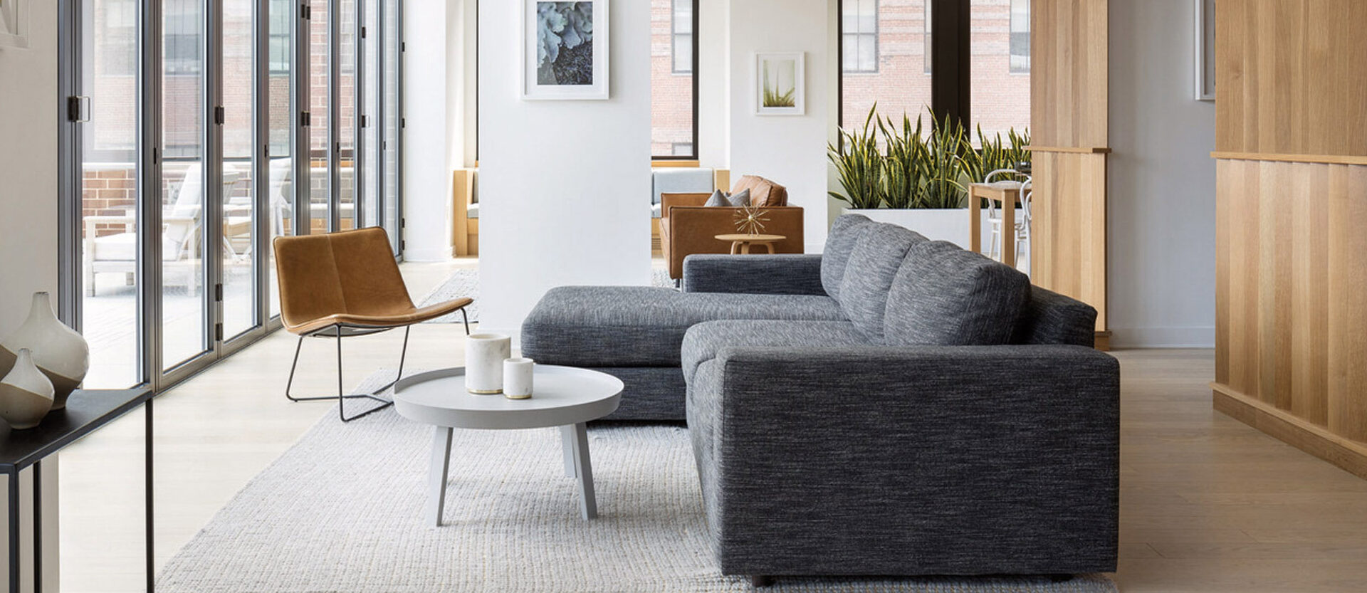 Bright, contemporary living space with minimalist design featuring clean lines. A dark grey sofa anchors the room against a large beige area rug, flanked by wooden bookshelves and modern leather armchairs. Natural light floods in through floor-to-ceiling windows, enhancing the warmth of the wooden accents.