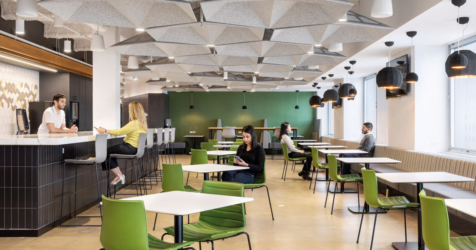 Modern office break room featuring acoustical ceiling panels, black pendant lighting, and lime green chairs paired with natural wood tables. People are casually interacting, reflecting functional design promoting productivity and social interaction.