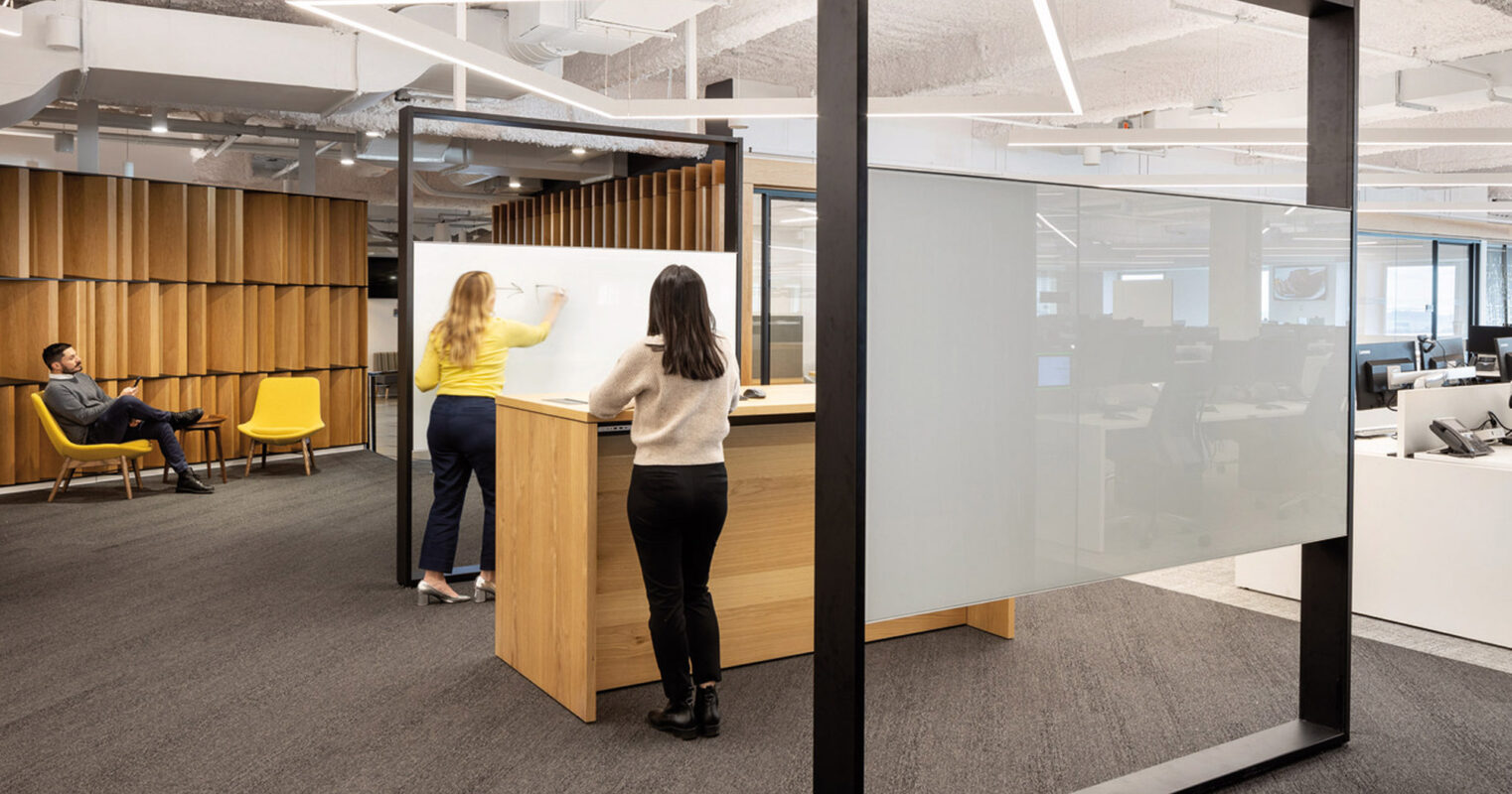 Open-plan office featuring a harmonious blend of natural wood panels and modern furnishings. Glass partitions provide transparency, while acoustic ceiling panels enhance comfort. Individuals engage in collaborative work in this well-lit, contemporary space.