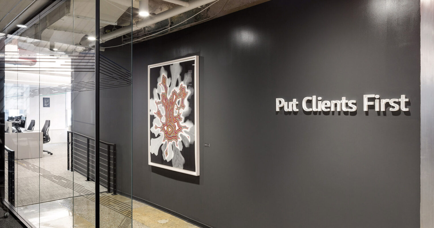 Modern office interior with industrial style featuring exposed ceiling and polished concrete floors. The focal point is a bold dark accent wall with motivational text "Put Clients First" beside framed abstract art, reflecting a professional yet creative workspace ambiance.
