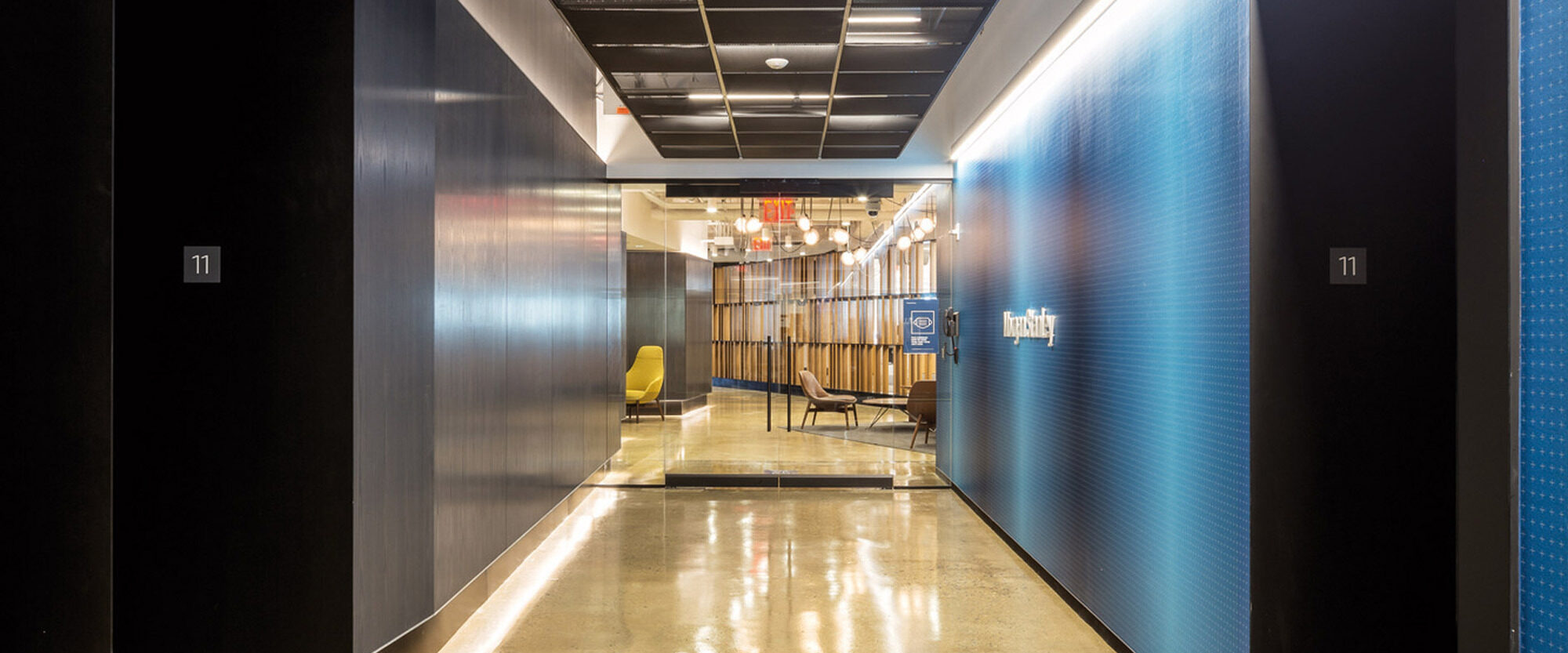 Modern office corridor with reflective polished concrete floors, contrasting blue-grey textured walls, and exposed ceiling structure showcasing a blend of industrial and contemporary design elements. Evening lighting adds warmth to the space.