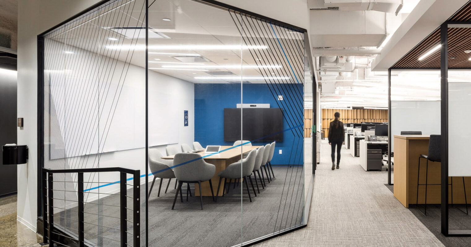 Modern office space featuring an open-plan layout with sleek glass partition walls. The meeting area includes a minimalist design with blue accent wall, complemented by grey carpeting and white ceiling exposing ductwork, revealing a blend of contemporary style with industrial elements.