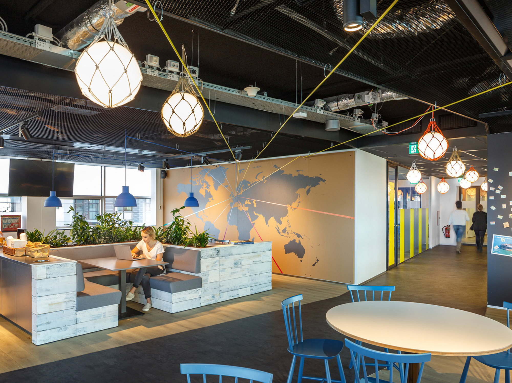 Modern office break area with eclectic hanging pendant lights, a world map mural on a white divider, and casual seating made from repurposed wooden pallets. The space features a mix of blue chairs, high ceilings with exposed ductwork, and an overall industrial chic vibe.