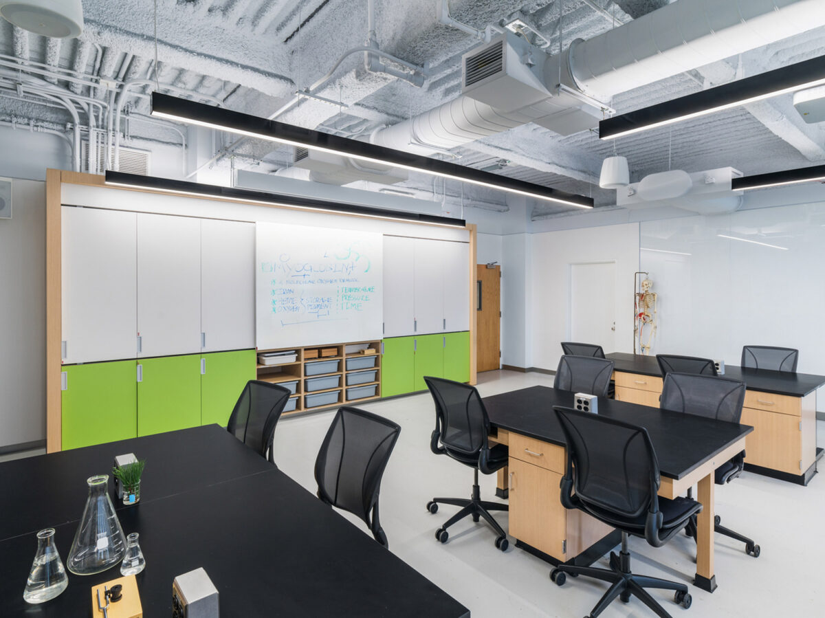Modern office meeting area featuring exposed ceiling infrastructure, whiteboard cabinetry, black rolling chairs, dark tables, and vibrant green accents. Natural light enhances the space's airy feel and complements the minimalist design approach.