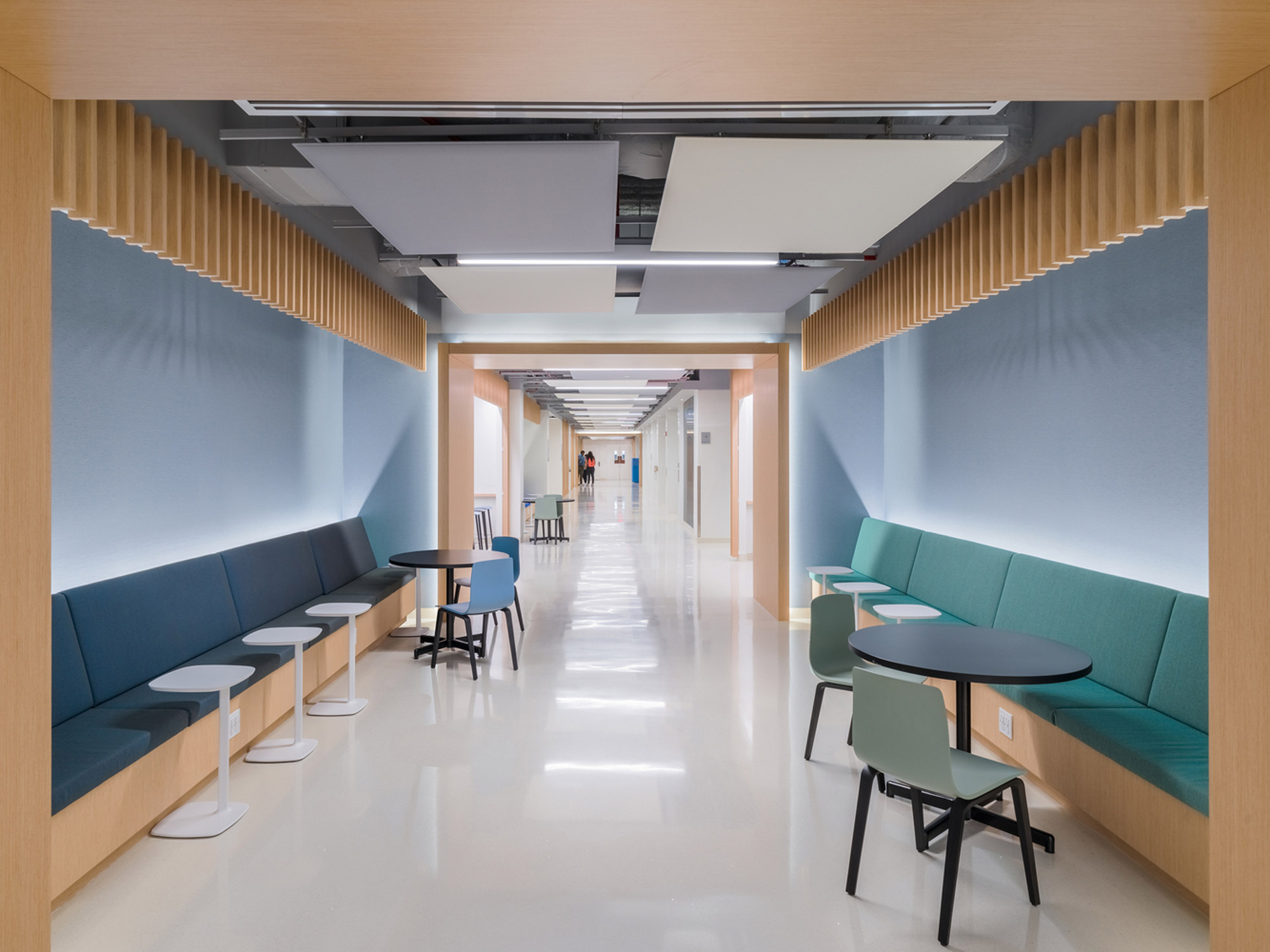 Modern corridor featuring sleek, high-gloss flooring that reflects the clean lines of recessed lighting. Neutral walls are accented by warm wooden slats, creating a tranquil environment. Strategically placed seating with teal and navy cushions provides comfort, complemented by minimalist round tables.