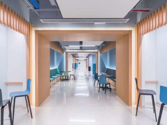 Modern corridor with clean lines, featuring wood-accented alcoves, teal and blue furnishing, and efficient LED lighting guiding the way. The design merges functionality with comforting aesthetics to ease navigation and enhance patient comfort.