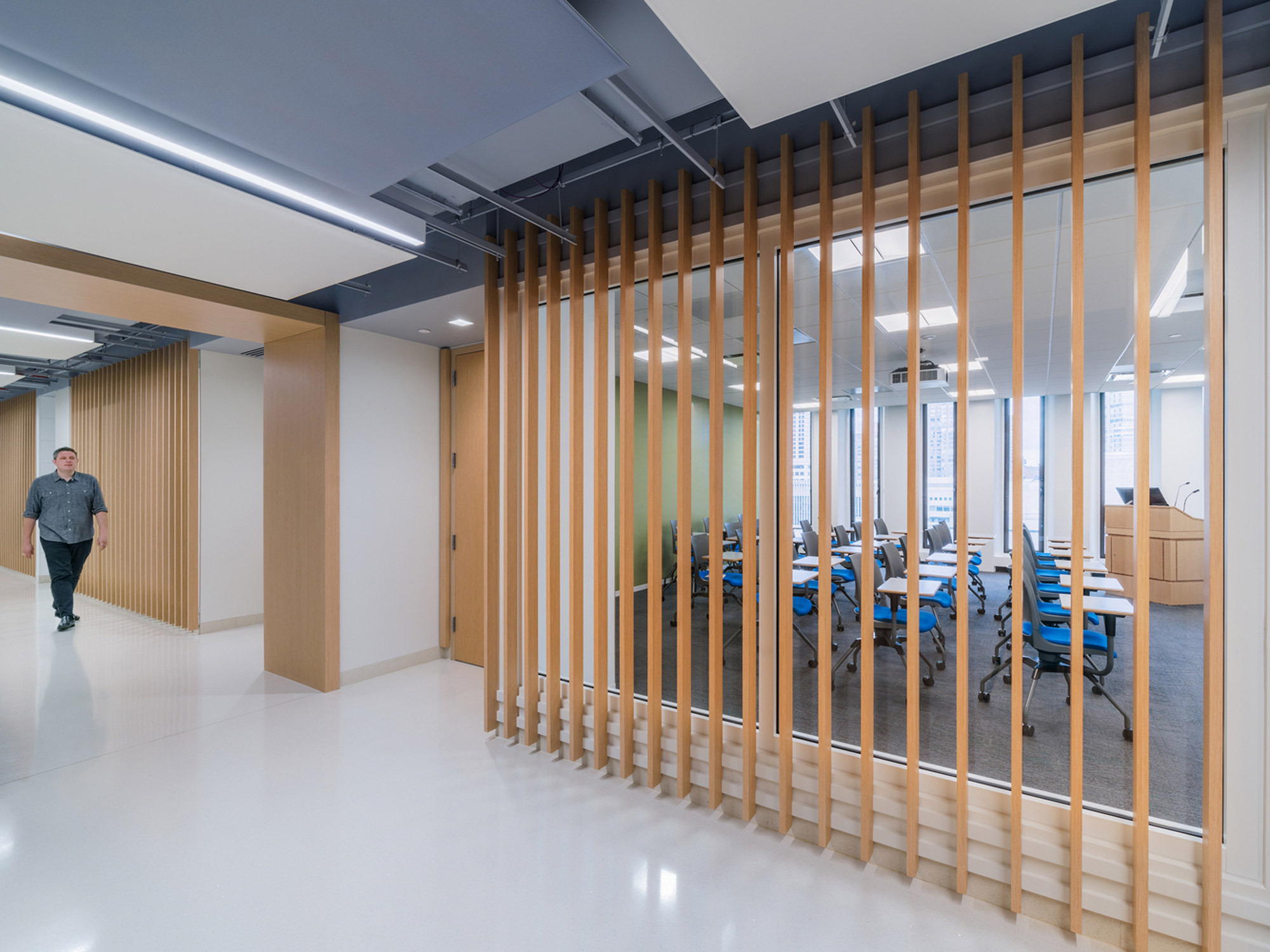 Modern office space showcasing a creative partition of vertical wooden slats, enhancing the flow between a walkway and a conference room with blue chairs. The design promotes transparency while offering a sense of separation, complemented by sleek lighting fixtures and a neutral color palette.