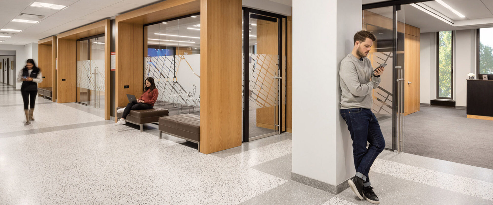 Modern office corridor featuring terrazzo flooring, neutral color palette, recessed lighting, and glass partition walls with etched geometric patterns. Wood paneling on doors adds a warm touch, juxtaposed with clean white columns and walls, offering a professional yet inviting atmosphere.