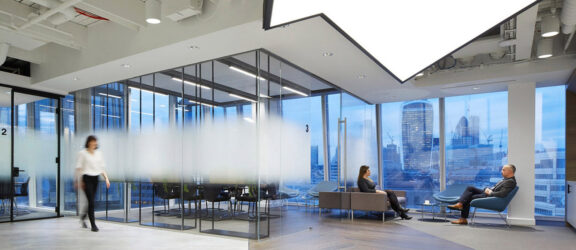 Modern office space featuring floor-to-ceiling glass partitions, sleek furnishings, and a neutral color palette. Ambient lighting complements the natural light flooding in from exterior windows, offering views of the urban skyline. Dynamic wood flooring contrasts with the structured ceiling design.