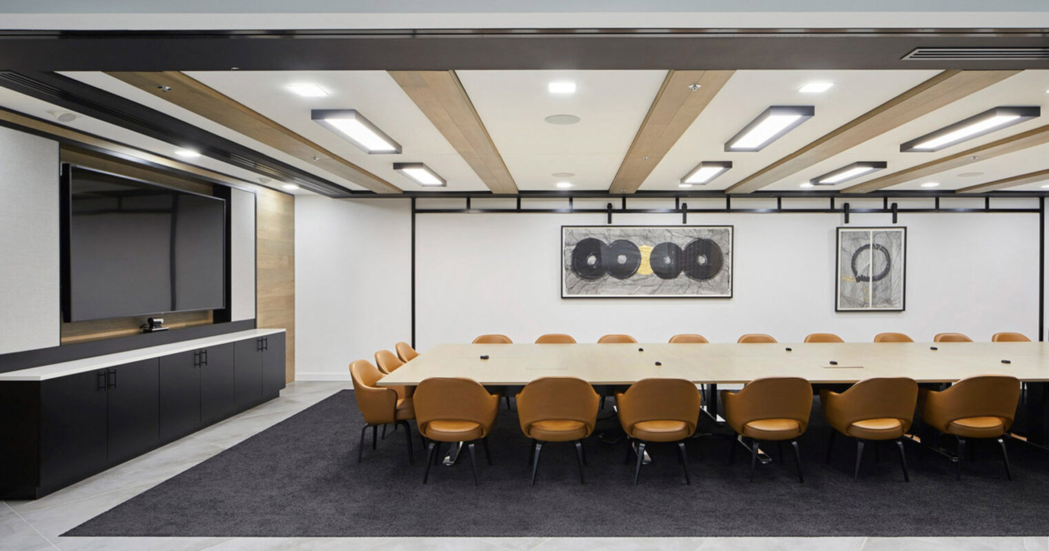 Modern conference room featuring a long wooden table surrounded by tan chairs, flanked by cabinetry and a large screen. Overhead, geometric light fixtures complement the linear wood-paneled ceiling, adding warmth to the neutral color palette. Two abstract monochrome artworks adorn the rear wall.
