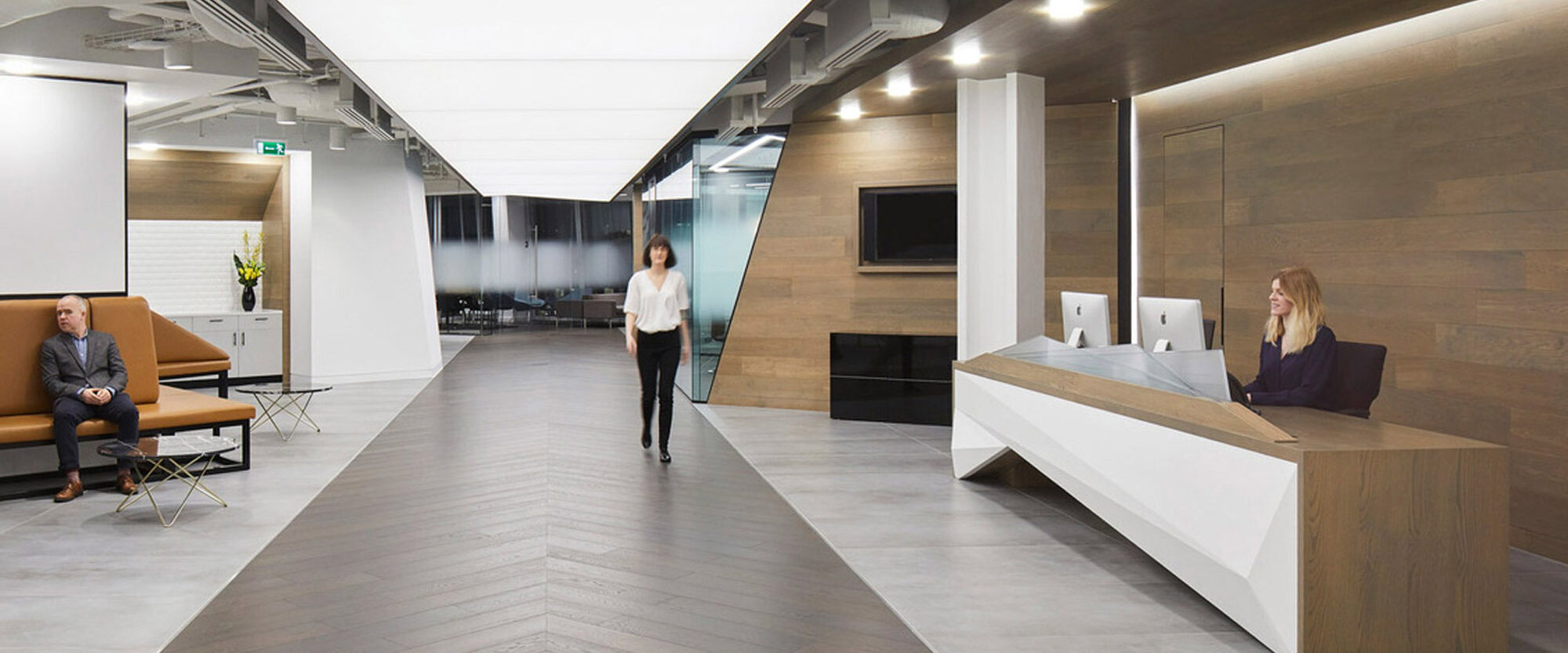 Modern office lobby with minimalist design, featuring a sleek white reception desk, wooden wall panels, herringbone floor pattern, and exposed ceiling, conveying an open and sophisticated space.