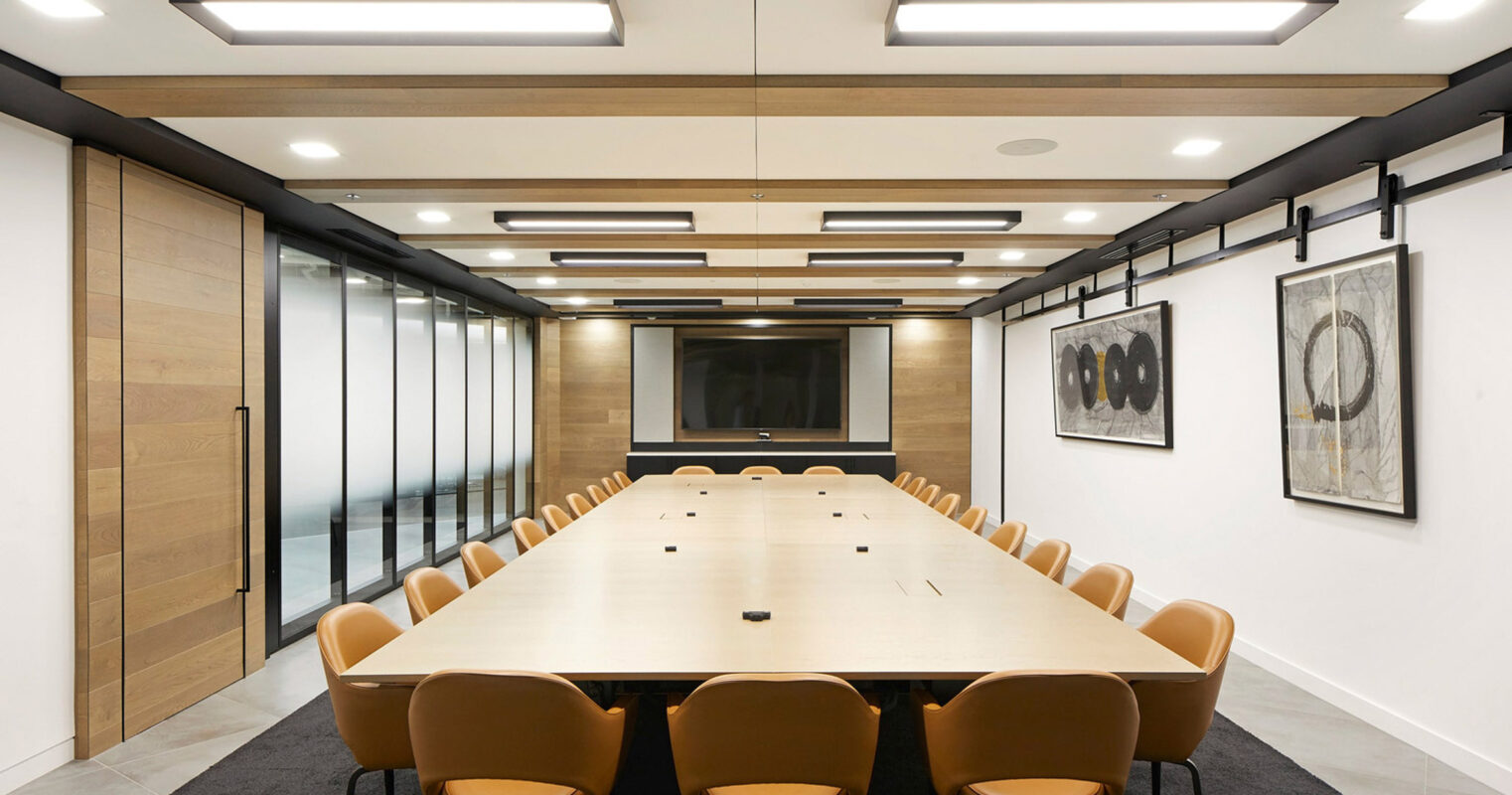 Modern conference room featuring a long rectangular table with plush orange chairs, wood paneling on walls, recessed and track lighting, accompanied by a flush-mounted video screen and framed art pieces.