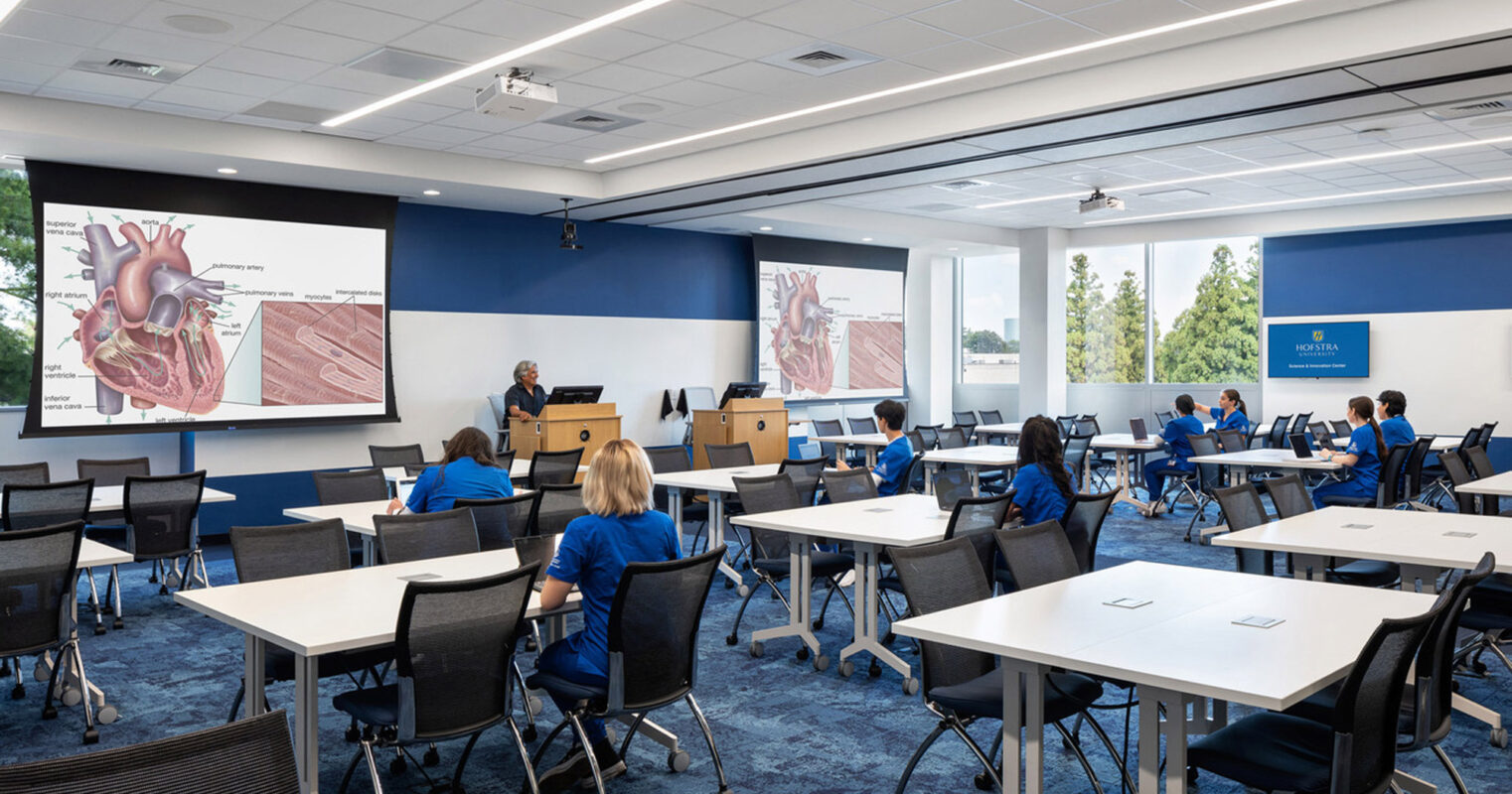 Spacious classroom featuring modern movable tables and chairs with a blue and white color scheme. The front wall displays a large projection screen, while ample natural light enhances the learning environment.