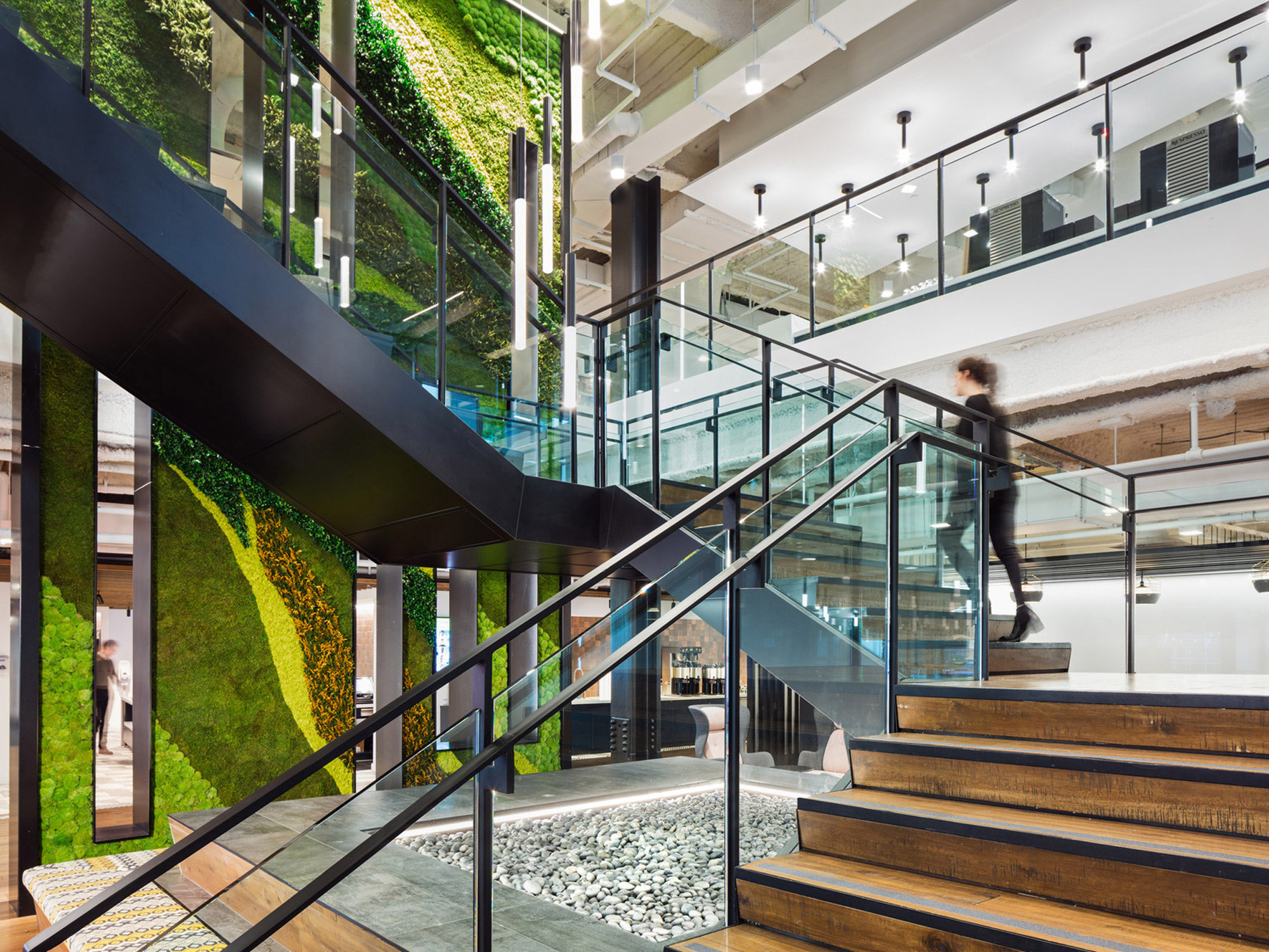 Modern interior with a biophilic design, featuring a spiral staircase with wooden steps, glass balustrades, and living green walls. The space includes concrete ceilings with exposed ductwork, black pendant lighting, and a pebble-filled atrium, emphasizing sustainability and connection to nature.