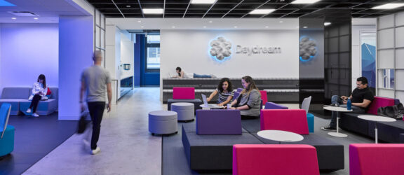 Modern office lounge featuring blue and magenta furniture with clean lines, accented by dynamic geometric carpet patterns. A boldly branded wall provides a backdrop to collaborative seating areas, emphasizing open-concept design and facilitating informal teamwork.