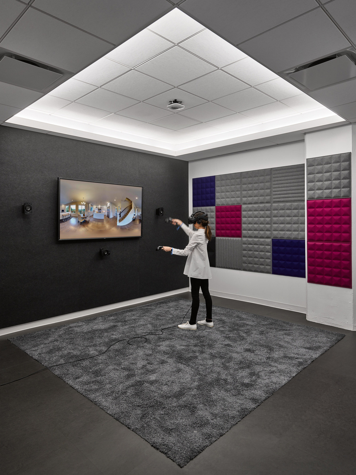 A modern interior features contrasting textured wall panels in grays and vibrant pinks, with a sleek digital display and a person gesturing towards the exhibit. The space is accentuated by a dynamic geometric ceiling design and a plush charcoal carpet, merging functionality with aesthetic appeal.