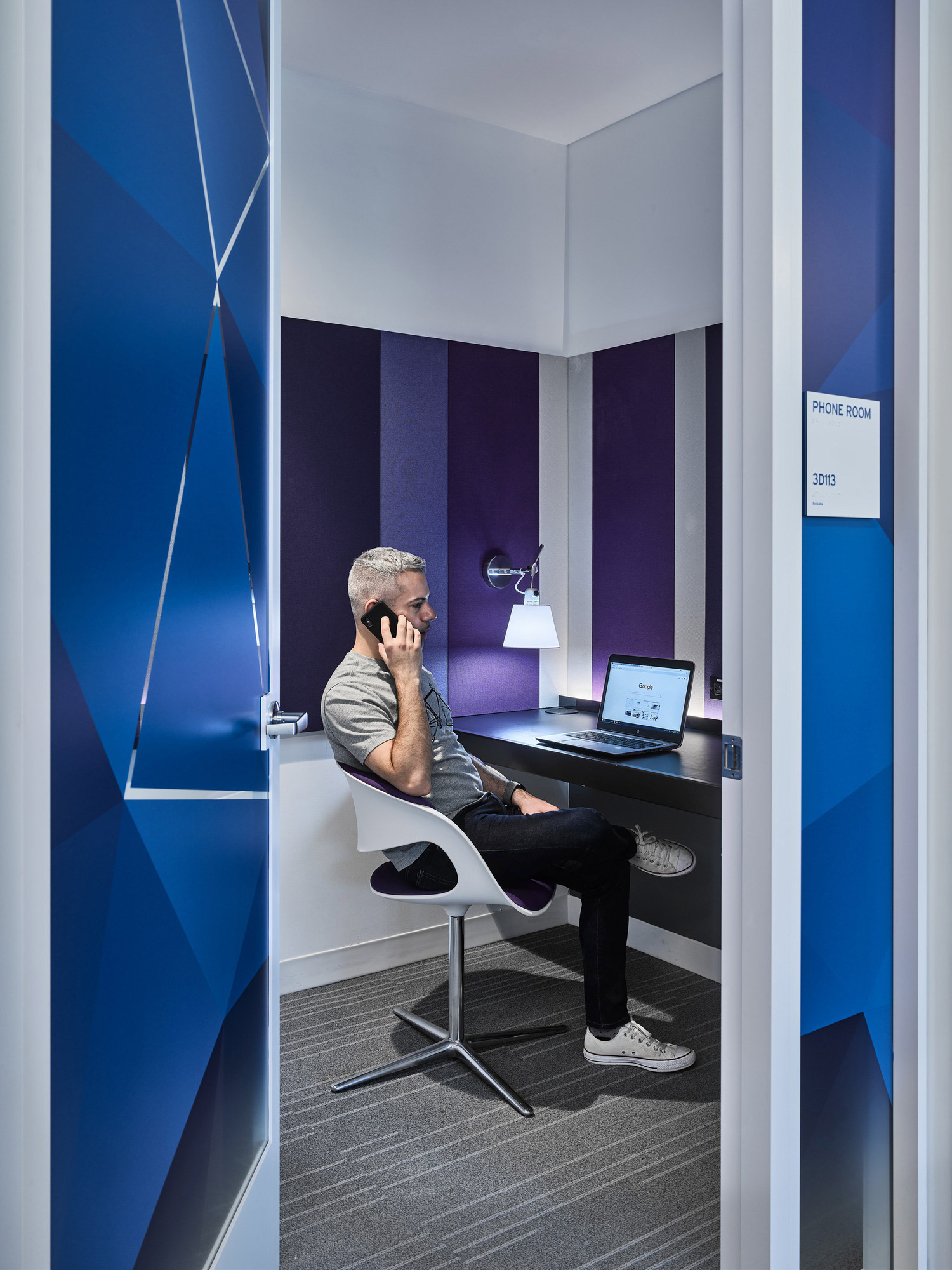 A compact office phone booth with geometric blue and white acoustic panels, featuring a built-in white desk with a mounted lamp and a man engaged in conversation on his mobile phone.