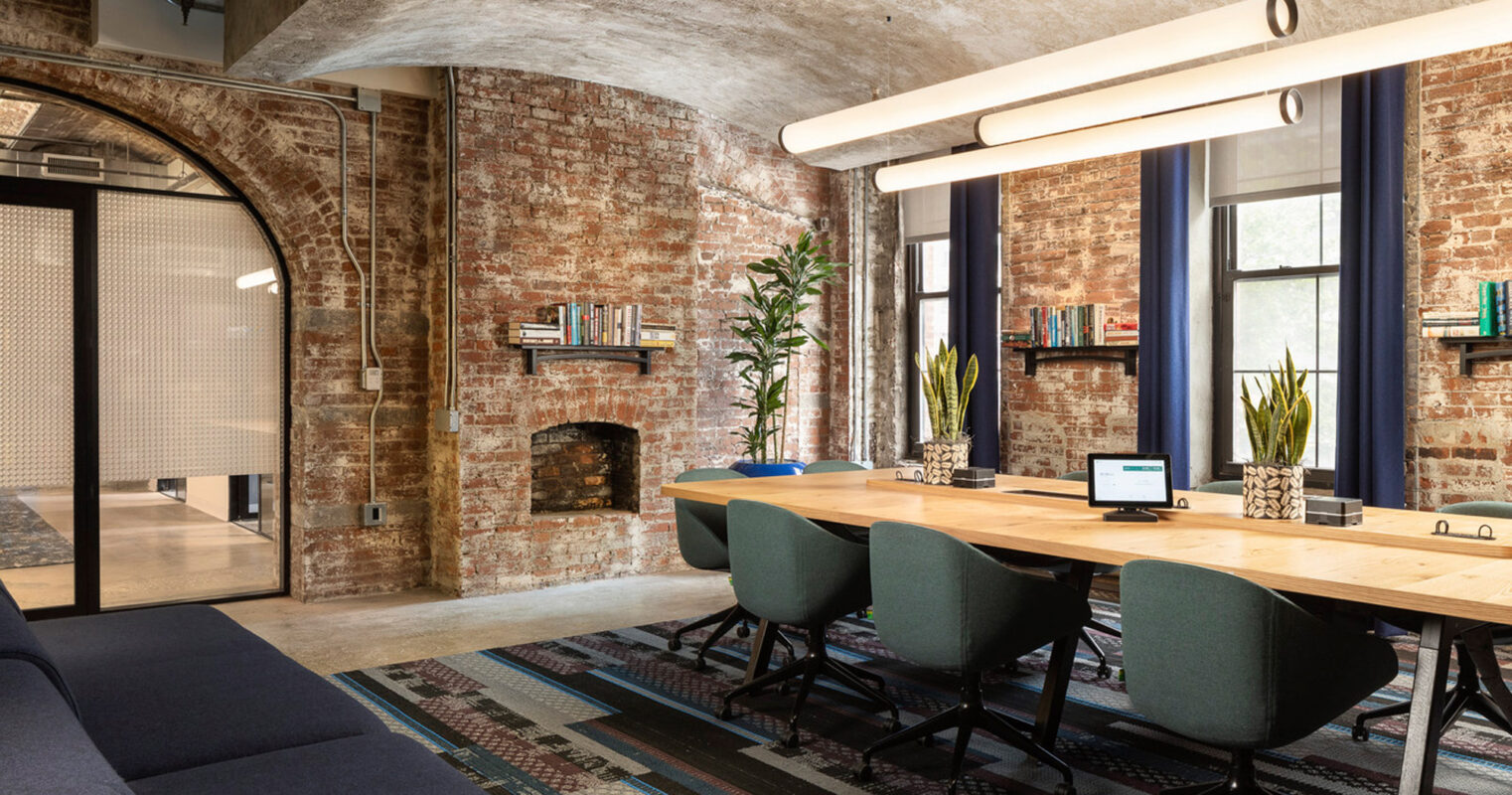 Modern office space blending industrial and contemporary design elements, featuring exposed brick walls, arched ceilings, and large windows complemented by sleek furnishings and patterned area rugs.