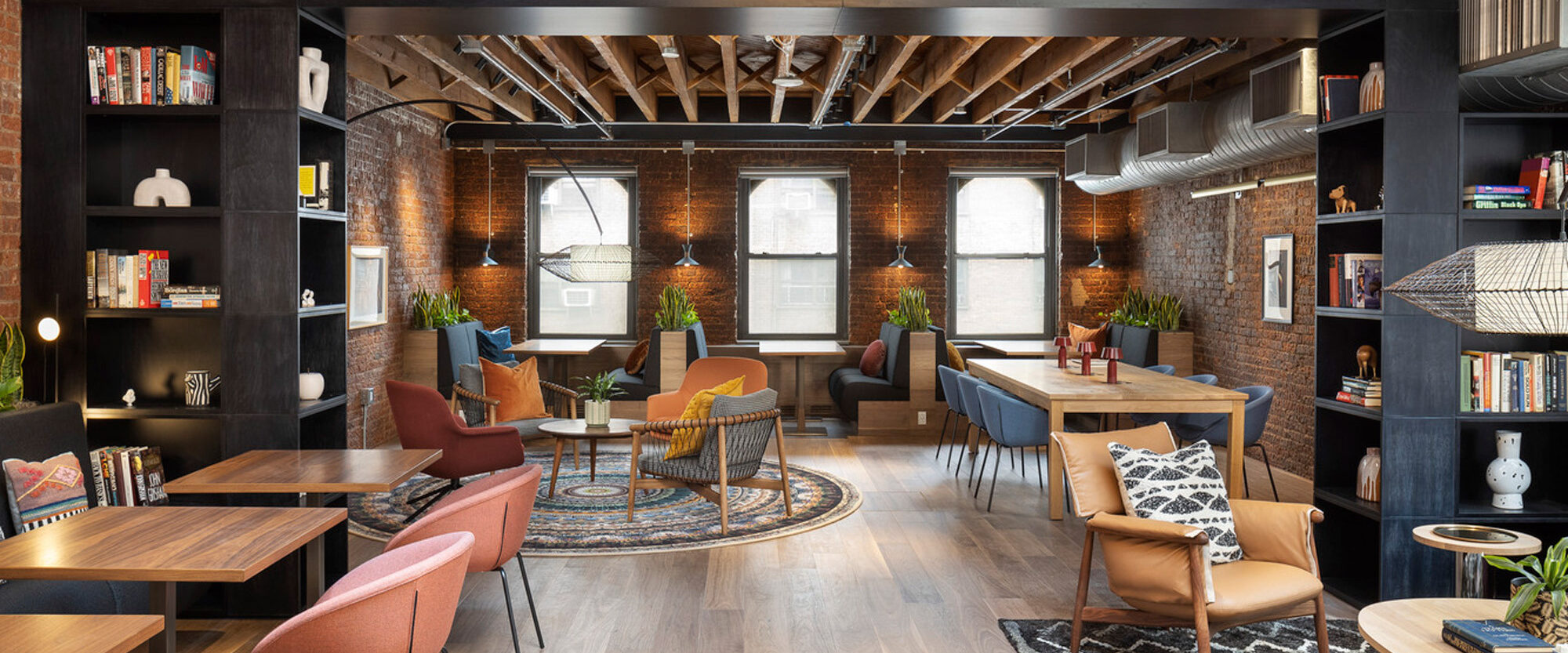 Spacious industrial loft featuring exposed brick walls and beam ceilings with contrasting modern furnishings in earth tones creates a warm, inviting atmosphere. Strategic lighting accentuates the bookshelves and seating areas, blending functionality with aesthetic appeal.