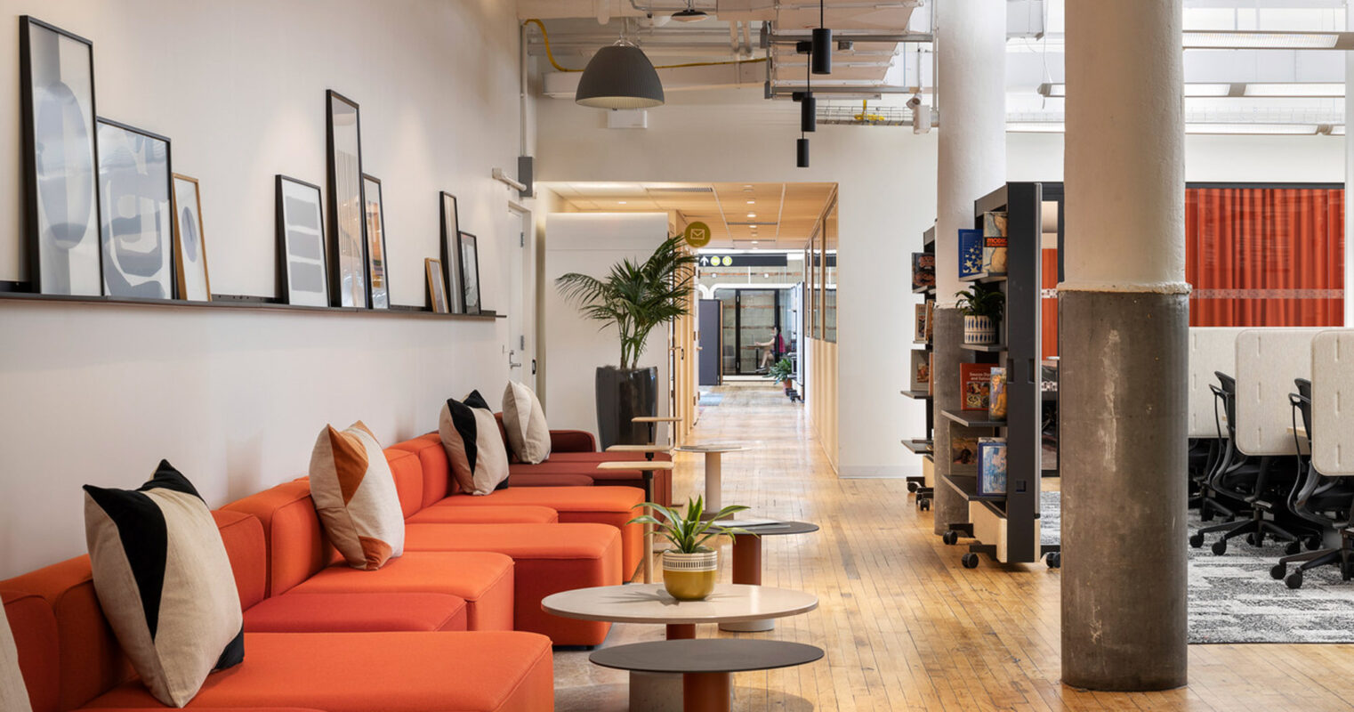 Modern open-plan office featuring a vibrant orange sectional sofa, eclectic accent cushions, and round coffee tables. Exposed beams and ductwork contrast with sleek furnishings and framed artwork, embodying industrial-chic design elements.