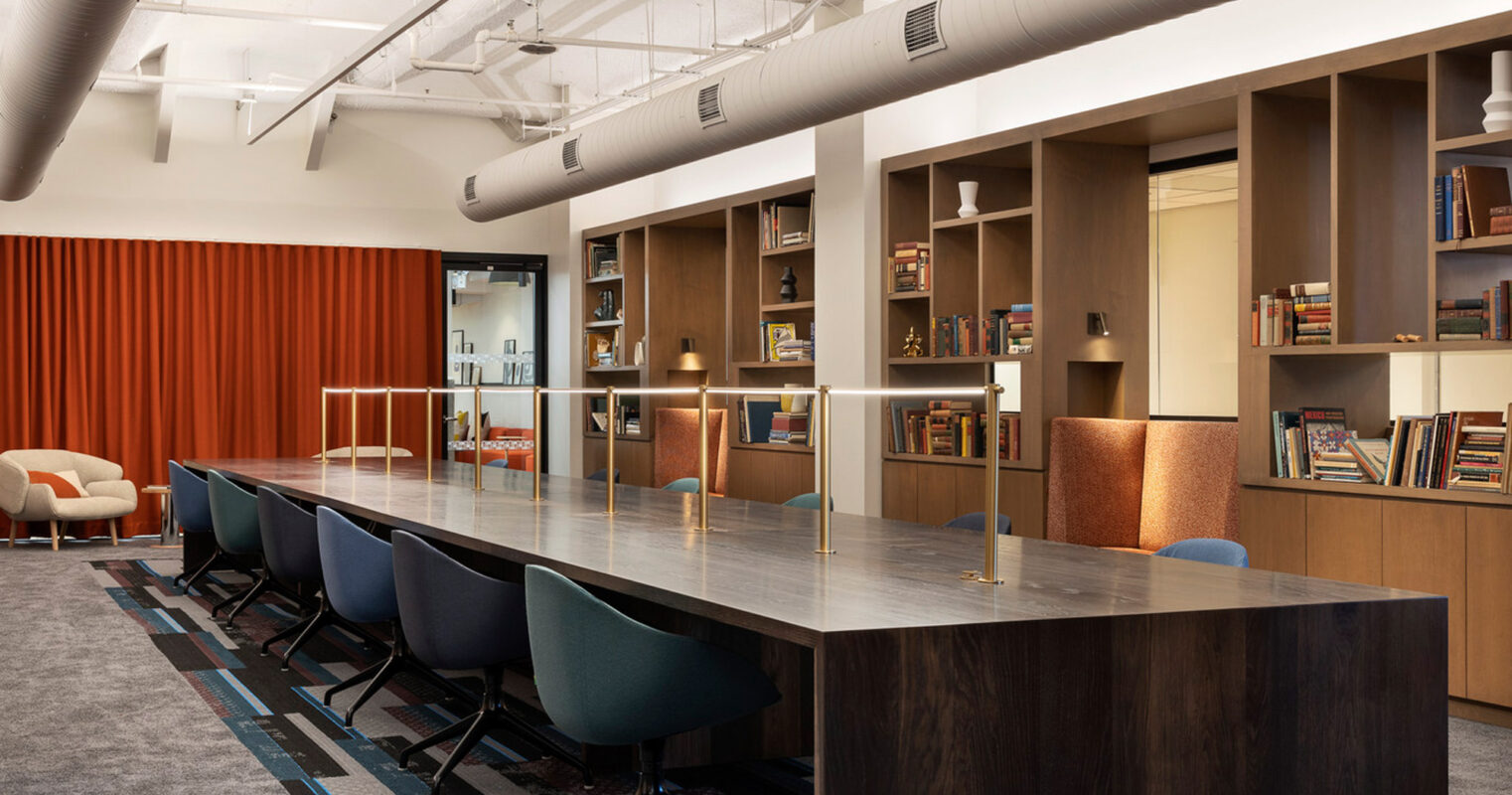 Modern office meeting area featuring exposed ceiling ductwork, a long dark wood table with teal chairs, a terracotta acoustic panel, and integrated shelving with decorative and informative literature, creating an industrial yet warm ambiance.