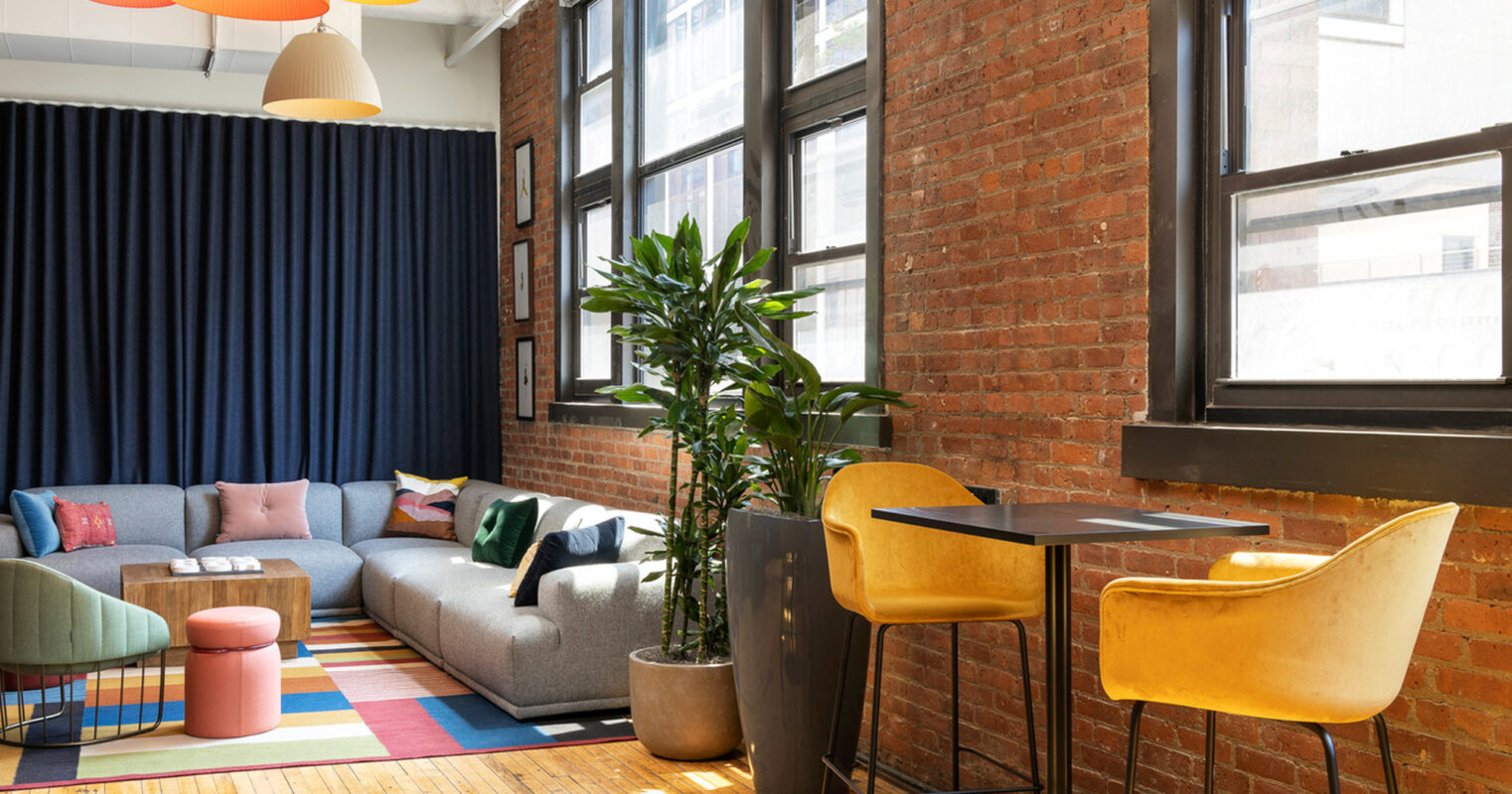 Modern loft living space with exposed brick walls and large windows. Varied colored pendant lighting hangs above a multi-textured seating area. A striped rug anchors the room, flanked by a tall green plant, leading to a cozy nook with bar-height chairs and a wooden table.