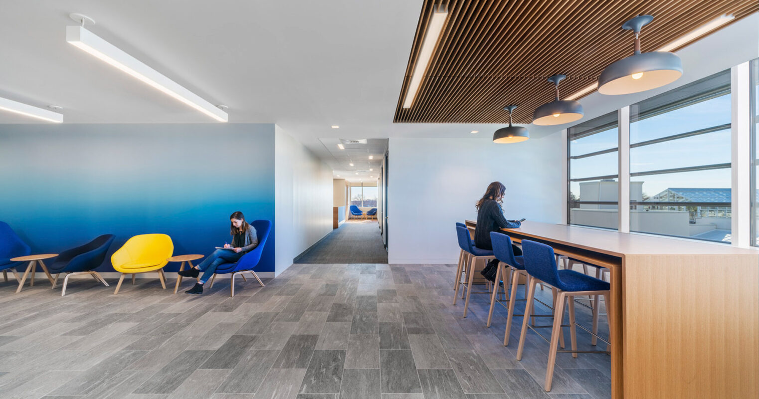 Contemporary office lounge with vibrant blue walls, contrasting yellow and blue furniture, and sleek, linear lighting fixtures. Wood accents in ceiling details and partitioning add warmth to the minimalist design, and floor-to-ceiling windows flood the space with natural light.
