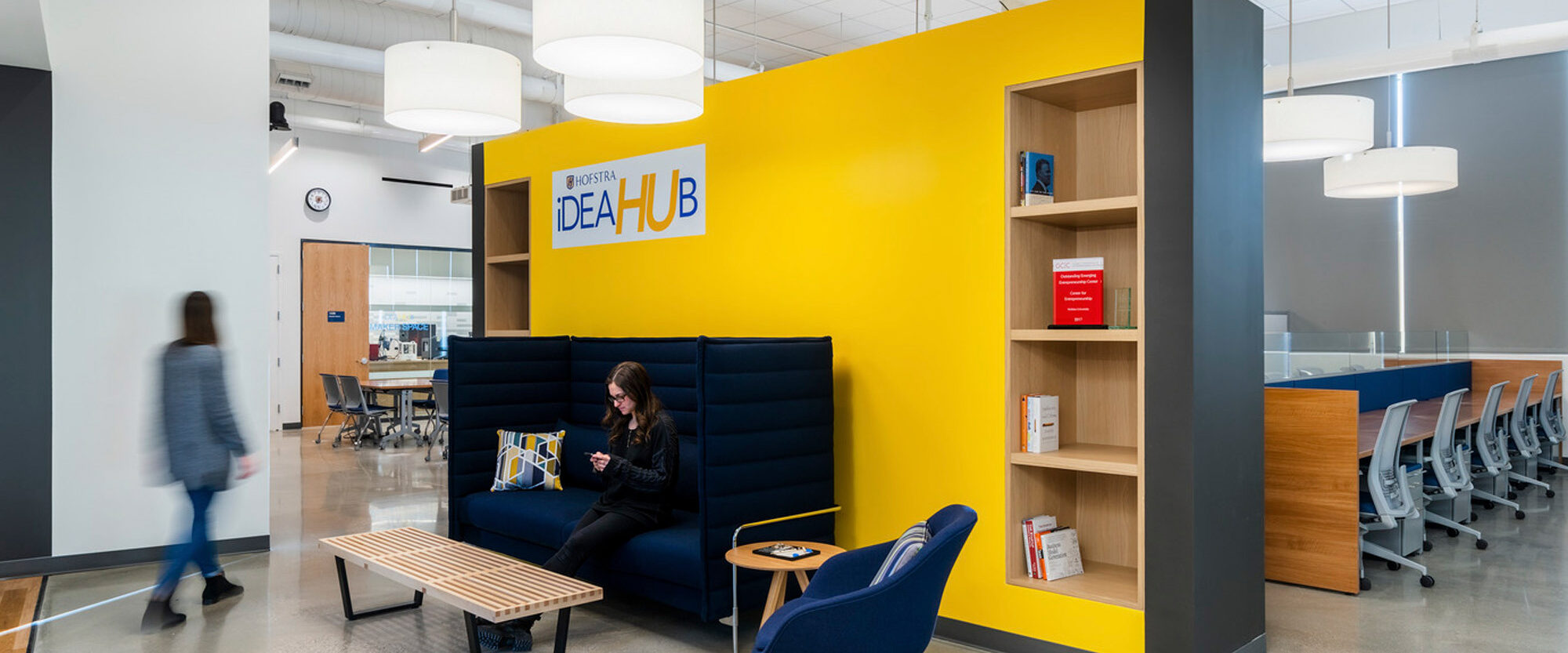 Open office space featuring a vibrant yellow accent wall with the text "IDEA HUB," an adjoining bookshelf, and modern pendant lighting. Individuals engage in work at communal tables, with lounge seating available for informal collaboration.