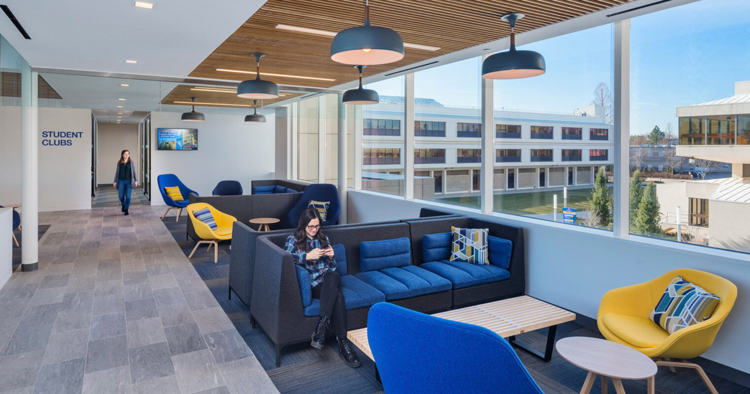 Modern open-plan student lounge featuring navy and yellow accent furniture, wood slat ceilings with pendant lighting, and expansive windows providing abundant natural light and a view of the surrounding campus.