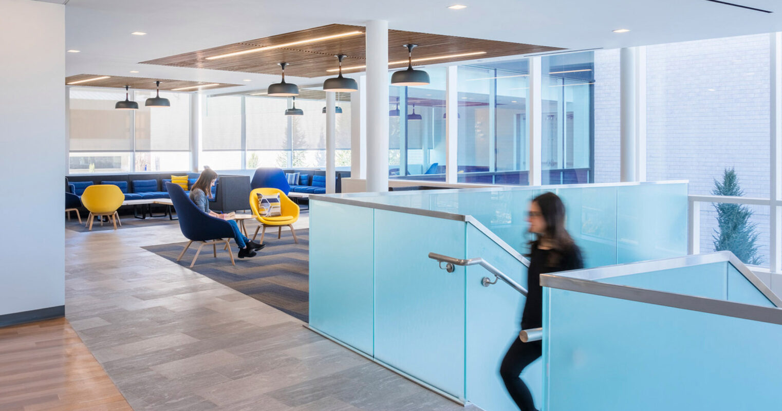 Modern office space with an open floor plan featuring clean lines, glass partitions, and a muted color palette with strategic accents of bright blue and yellow. Pendant lighting and contemporary furnishings create a sophisticated yet inviting work environment.