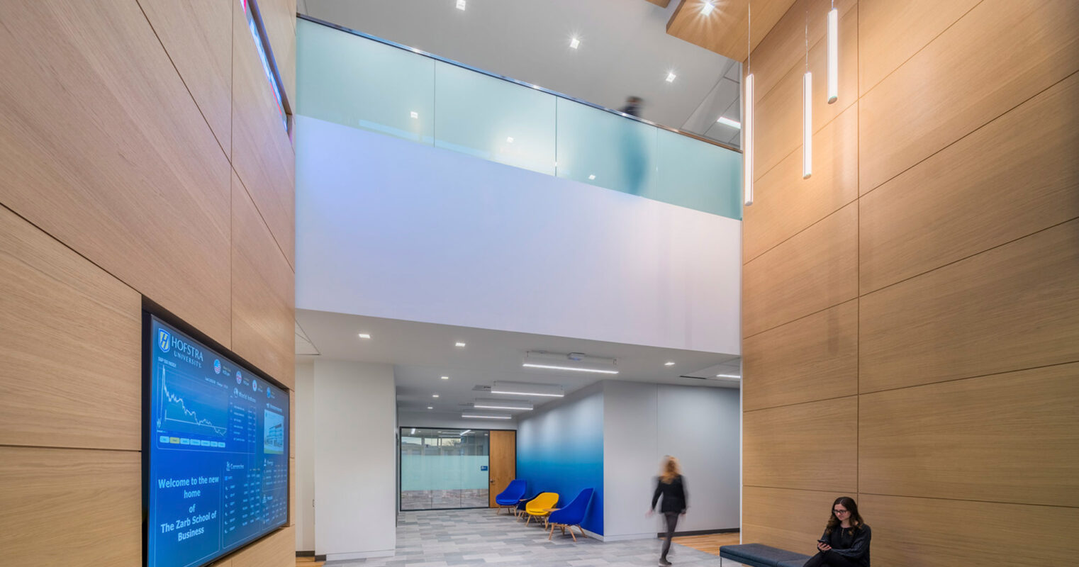 Modern office lobby featuring a warm wood-paneled wall, geometrically patterned floor, sleek hanging lights, and a digital information display. The space is illuminated by natural light from clerestory windows, enhancing the open-plan design with a blue accent chair adding a pop of color.