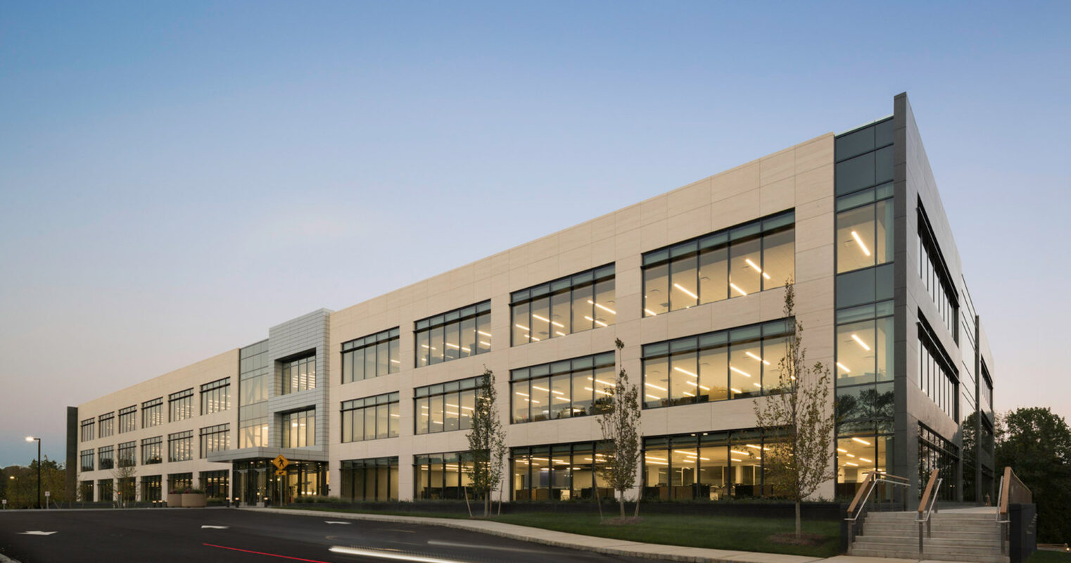 Modern office building exterior at dusk, featuring clean lines, expansive glass windows, and a mix of white paneling. The design emphasizes transparency and open spaces, highlighted by the evening's soft ambient light.