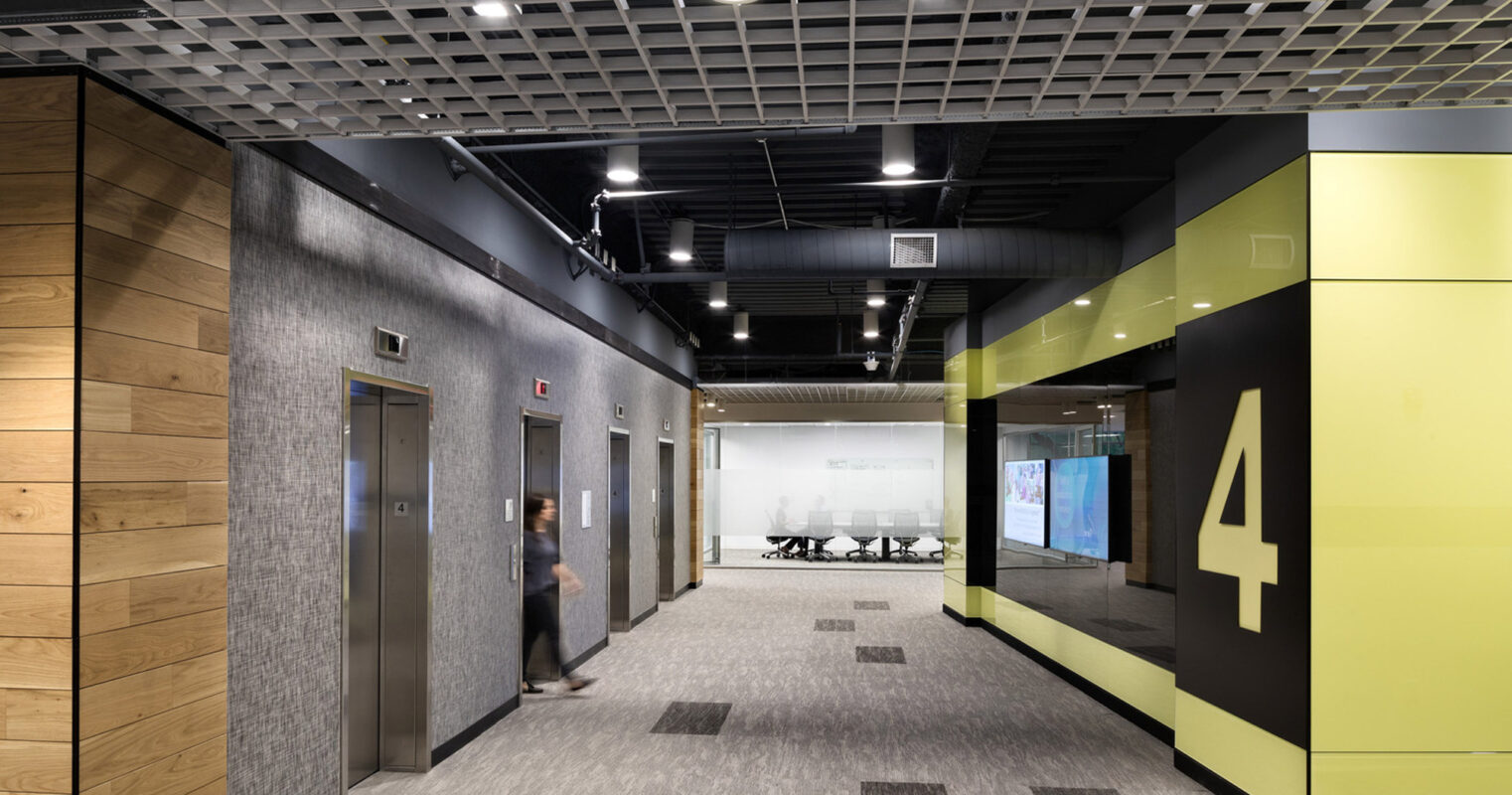 Modern office corridor with textured wood walls and geometric carpet. Black ceiling grid contrasts with recessed lighting above, while lime green accents energize the space. Transparent glass partitions reveal meeting room interiors, enhancing openness. Large numerals denote room numbers, guiding navigation.