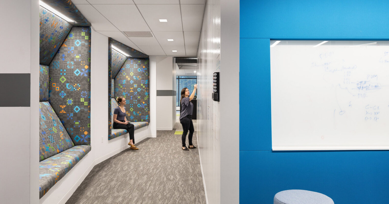 Modern office corridor featuring vibrant geometric-patterned wall upholstery, recessed lighting, and sleek gray flooring. Two professionals engage near a whiteboard reflecting collaborative work in a contemporary workspace.