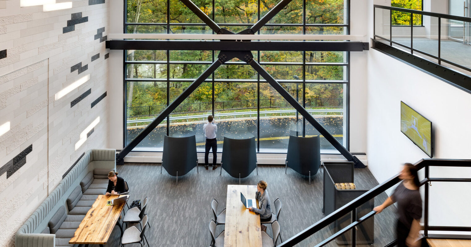 Modern office interior with high ceilings and large picture windows showcasing an autumnal tree panorama. Natural light illuminates the minimalist décor, contrasting the industrial-style black beams and railings. Open-plan seating area features wood tables and gray upholstered benches, fostering a collaborative workspace.