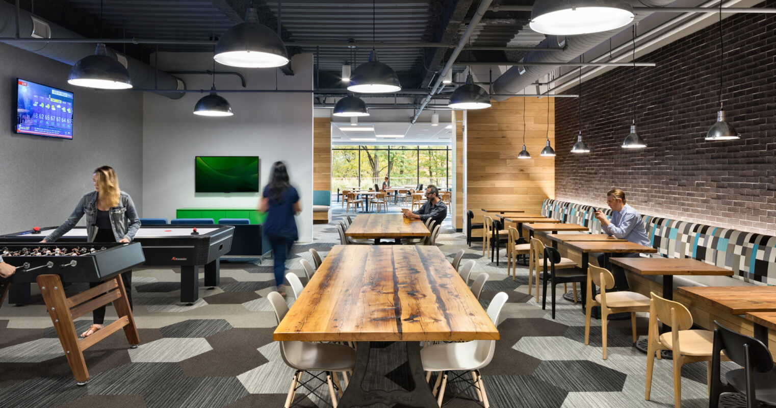 Modern office break room featuring a rustic wooden communal table, individual tables with chairs, pendant lighting, a foosball table, and a relaxed, collaborative atmosphere. An exposed ceiling and large windows create an airy, well-lit space.