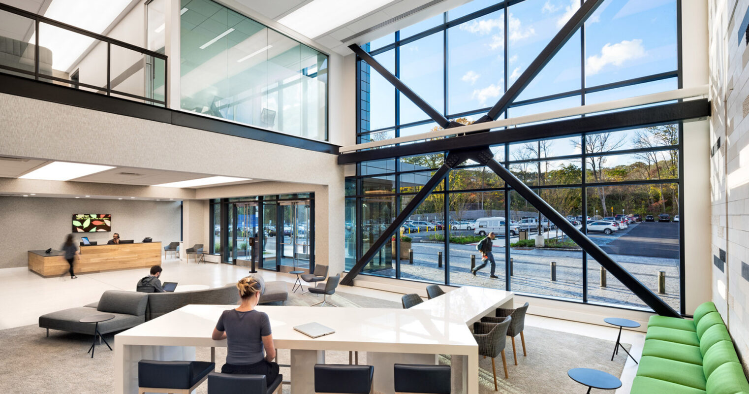 Spacious modern lobby with expansive glass walls, geometric black support beams, and abundant natural light. A reception desk pairs with minimalist furniture, featuring a neutral color palette with green accent seating, promoting a sleek, open-plan corporate environment.