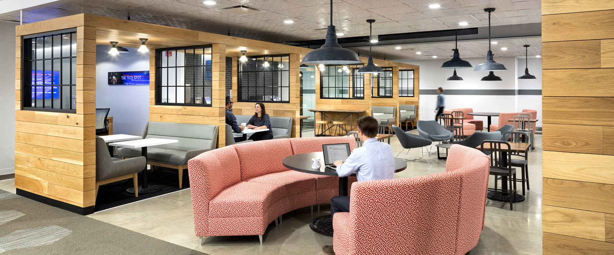 Modern office breakroom featuring a cohesive blend of natural wood structures and industrial-style pendant lighting. Curved coral sofas provide a pop of color, complemented by gray lounge chairs and bar-style seating, creating a multifunctional, inviting space for collaboration and relaxation.