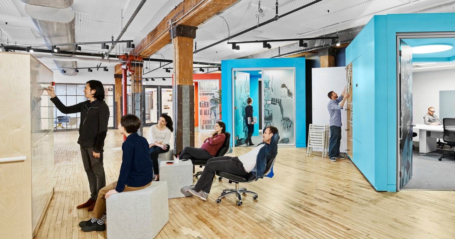 Open-plan office space blending modern and industrial elements, featuring exposed brick and wooden beams, vibrant teal office pods with frosted glass, hardwood floors, and a mix of seated and standing workspace options. Employees actively engage in collaborative and individual tasks.